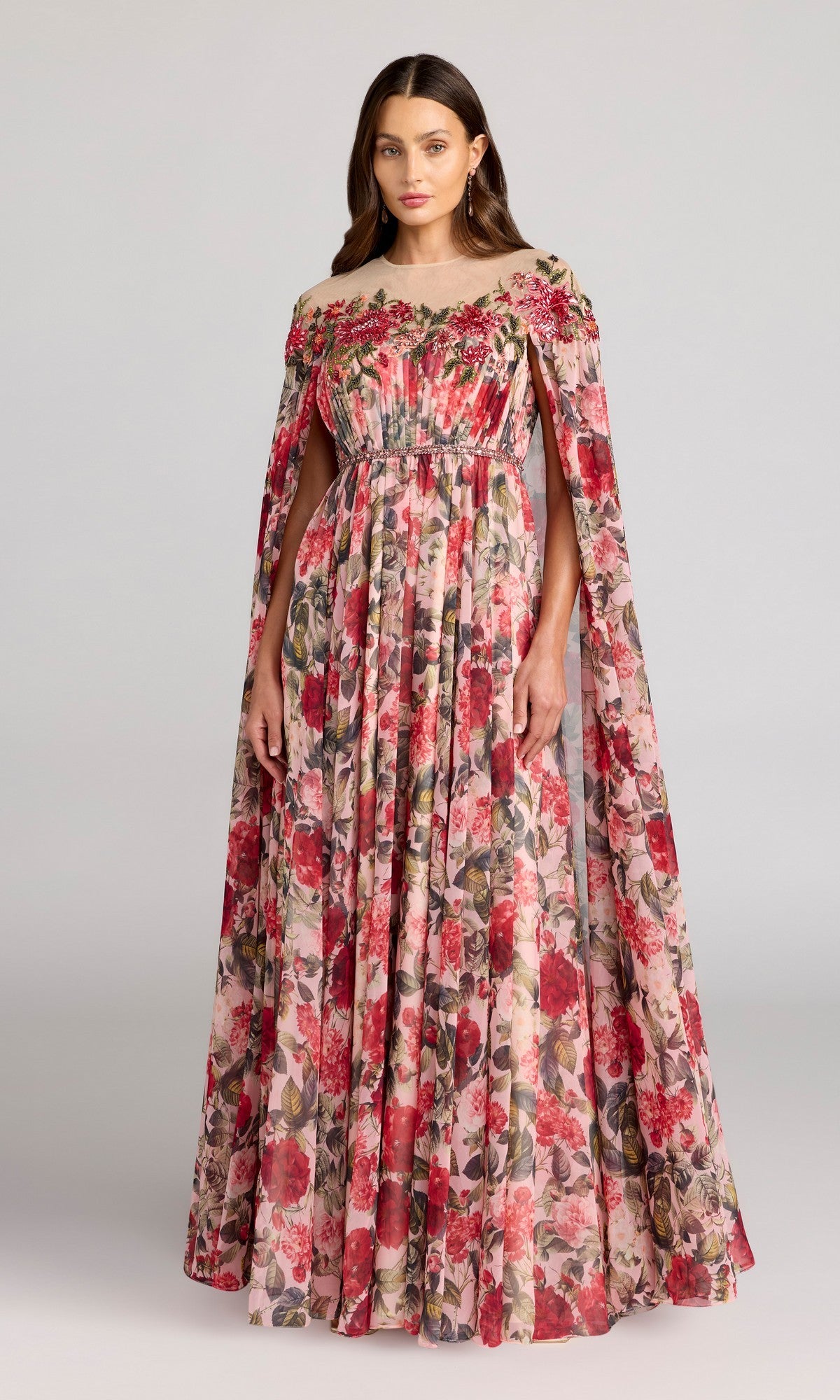 A woman models the Mac Duggal 11907 long formal dress in Rose Multi, featuring a floor-length chiffon design with elegant cape sleeves. The gown showcases a gathered bodice and delicate floral prints in shades of red, pink, and green, beautifully set against a light gray background.